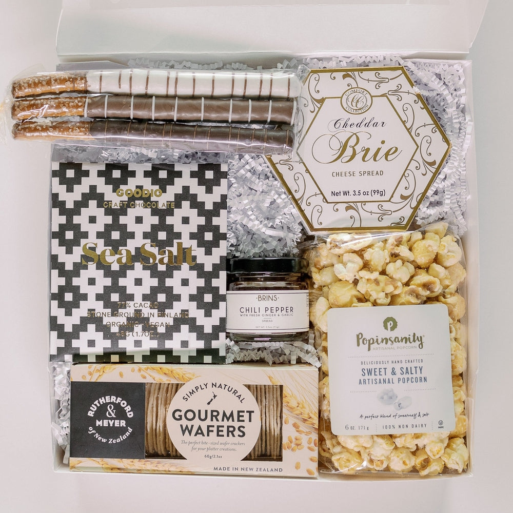 Photograph of a gourmet gift box containing artisan snacks featuring striped chocolate-covered pretzels, a gold-trimmed hexagonal cheese spread container, and a black-and-white geometric patterned Goodio chocolate bar on white crinkle paper bedding