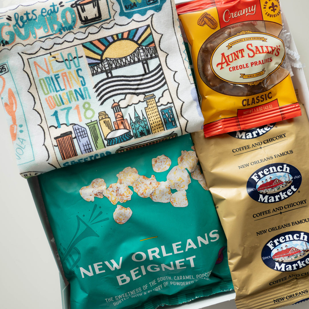 Photograph of a New Orleans themed gift box featuring an illustrated tea towel depicting the St. Louis Cathedral and Superdome landmarks alongside an orange pack of Aunt Sally’s Creole Pralines and a vibrant teal beignet snack bag
