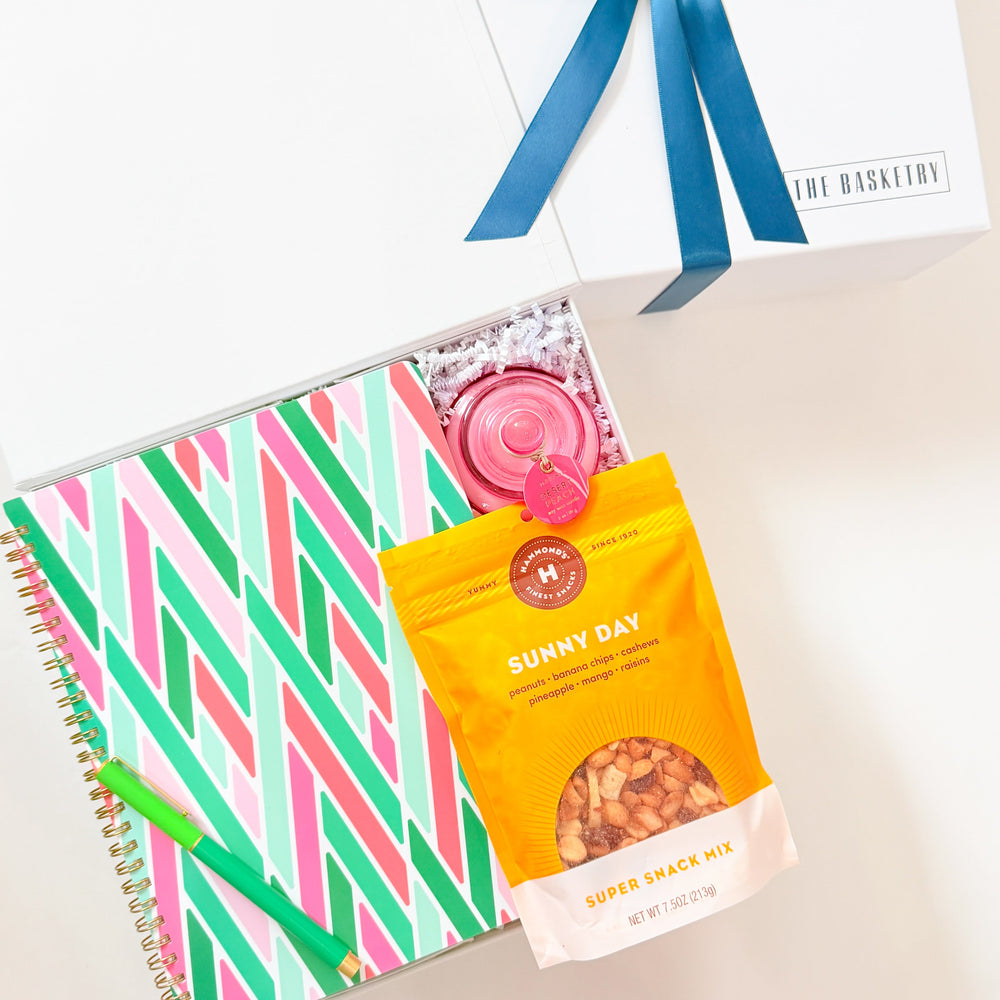 Spiral-bound notebook with colorful striped design, a bag of Sunny Day snack mix, and a card with a blue ribbon on a light background.