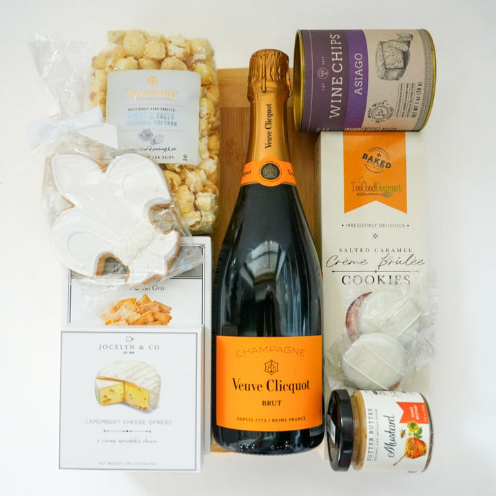 Digital photograph of a luxury gourmet gift assortment on a white surface featuring a bottle of Veuve Clicquot Brut Champagne accompanied by a white fleur-de-lis iced cookie, a purple tin of Asiago Wine Chips, and artisanal Camembert cheese spread