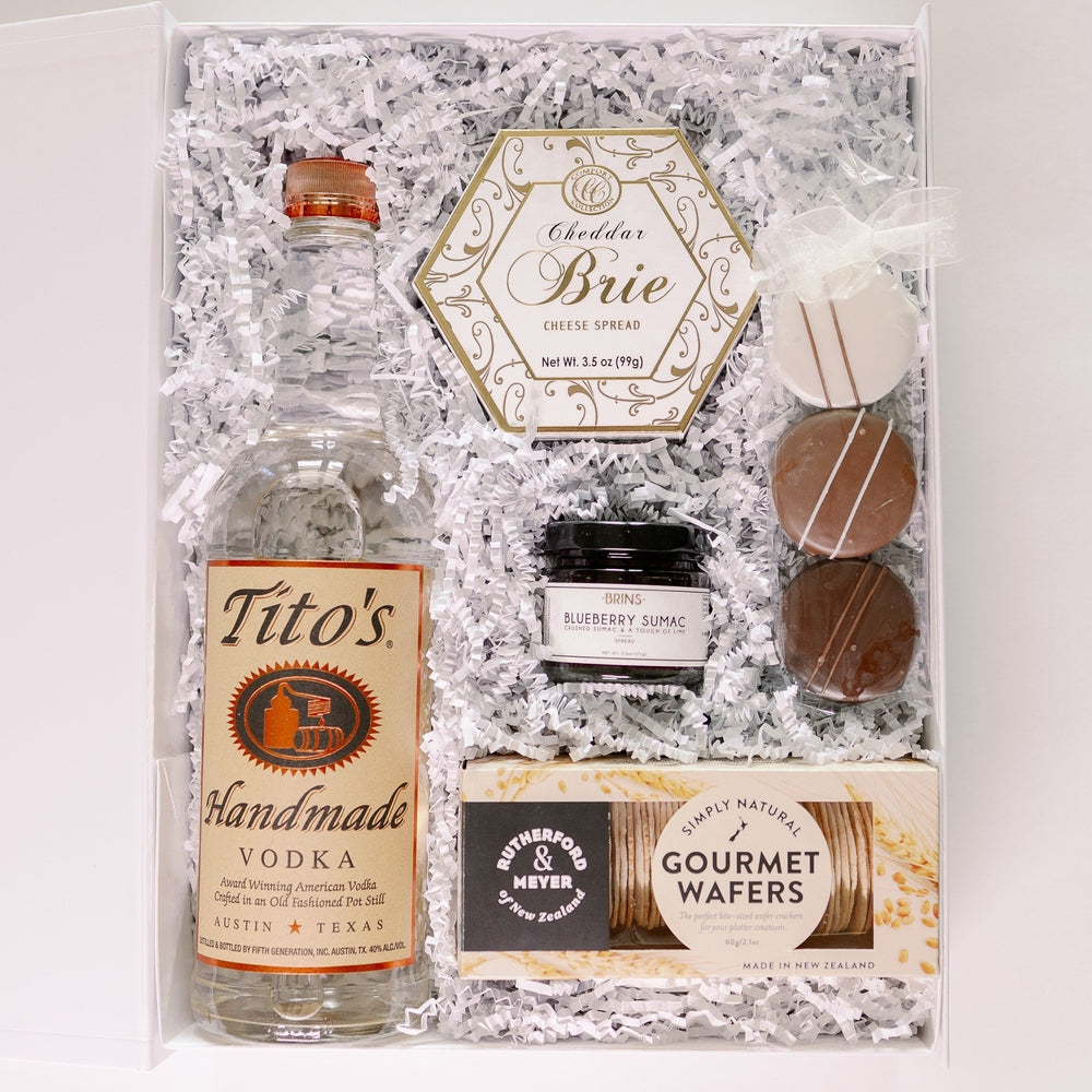 Digital photograph of a gourmet gift basket featuring a bottle of Tito's Handmade Vodka from Austin, gold-filigreed Cheddar Brie cheese spread, and New Zealand gourmet wafers nestled in white shredded paper with striped chocolate truffles