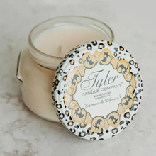 Product photograph of a Tyler Candle Company scented candle on a light marble surface featuring an open glass jar with a decorative leopard print lid, ornate gold filigree, and smooth cream-colored wax