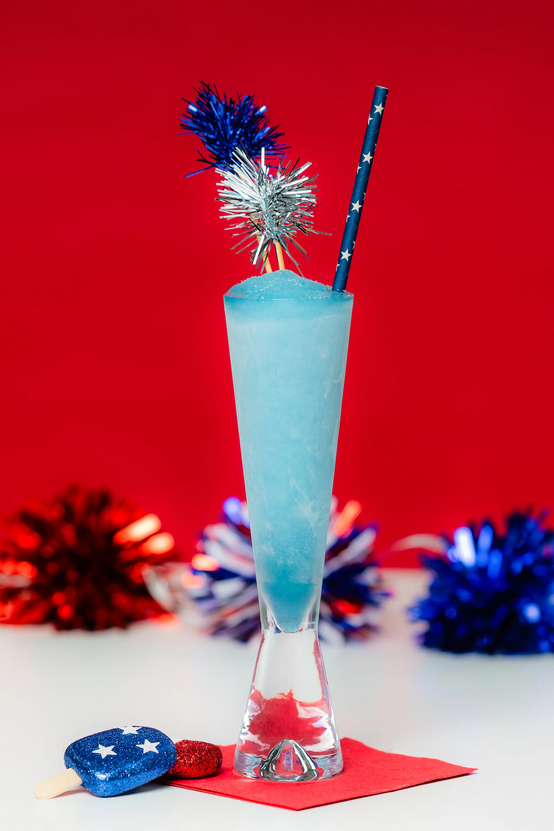 Photograph of a vibrant light blue frozen cocktail in a tall glass featuring a blue star-patterned straw, shimmering tinsel picks, and a glittery patriotic popsicle accessory celebrating Independence Day against a bold red backdrop