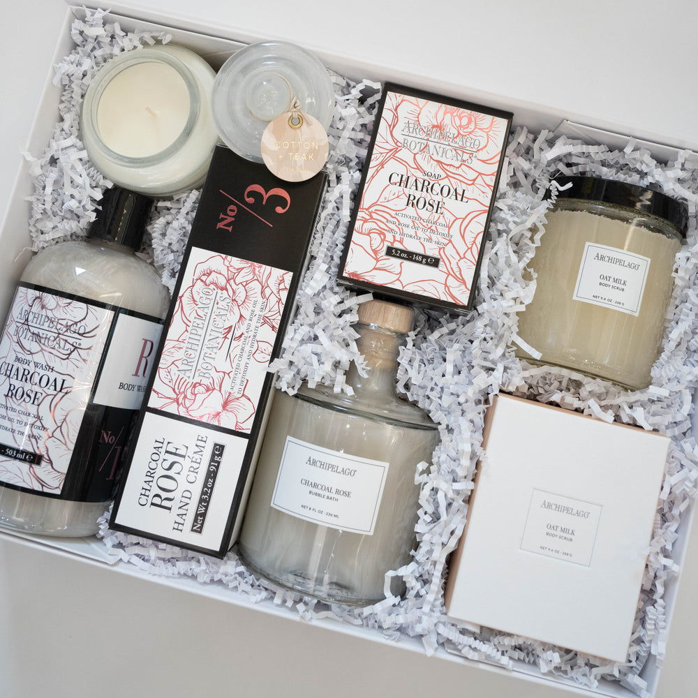 Digital photograph of an Archipelago Botanicals luxury gift set in a white box featuring Charcoal Rose and Oat Milk products nestled in white crinkle paper with pink floral packaging and a cork-stoppered glass bottle