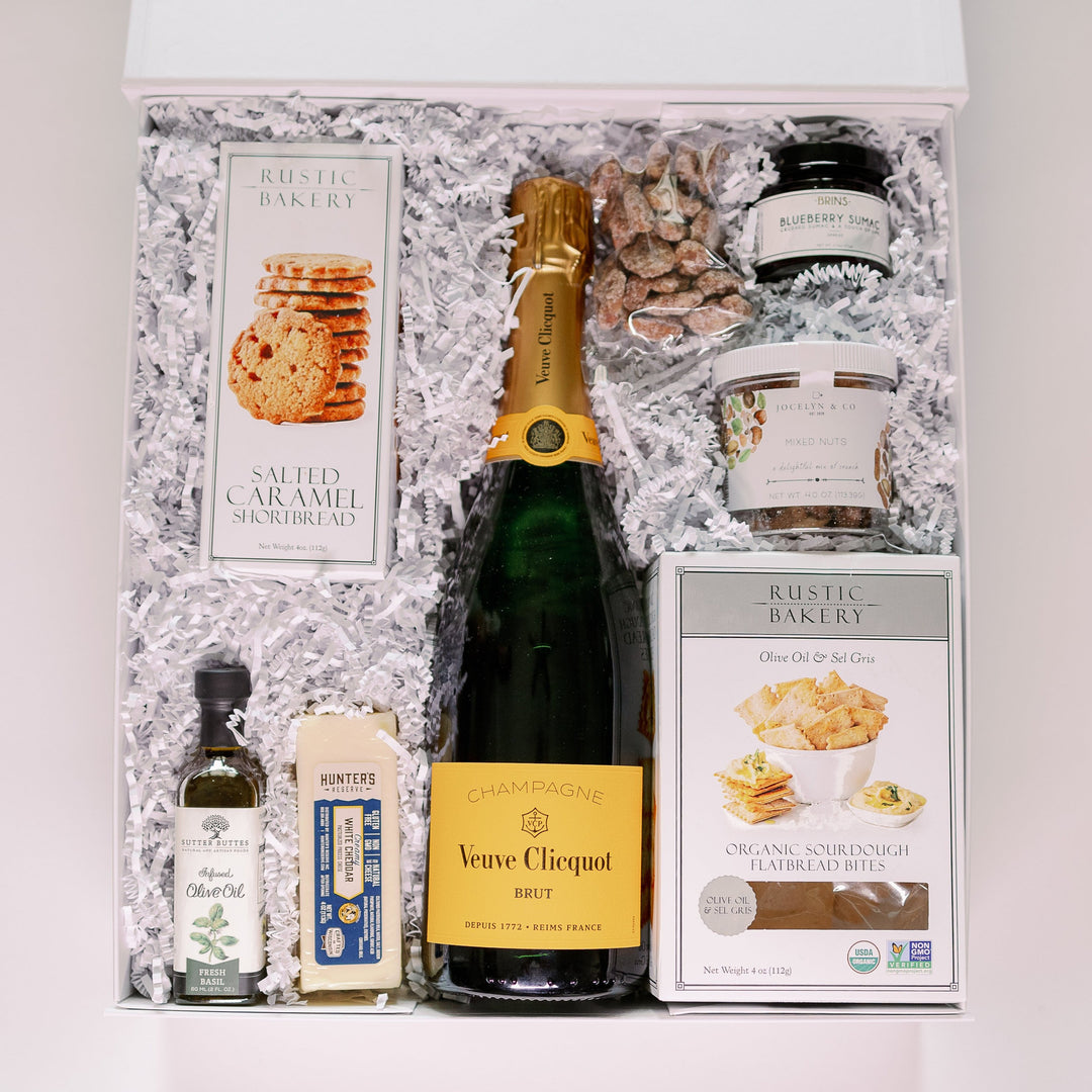 Digital photograph of a luxury gourmet gift hamper featuring a bottle of Veuve Clicquot Brut Champagne with its signature yellow label and Rustic Bakery treats nestled in white shredded paper alongside artisanal white cheddar and basil infused olive oil