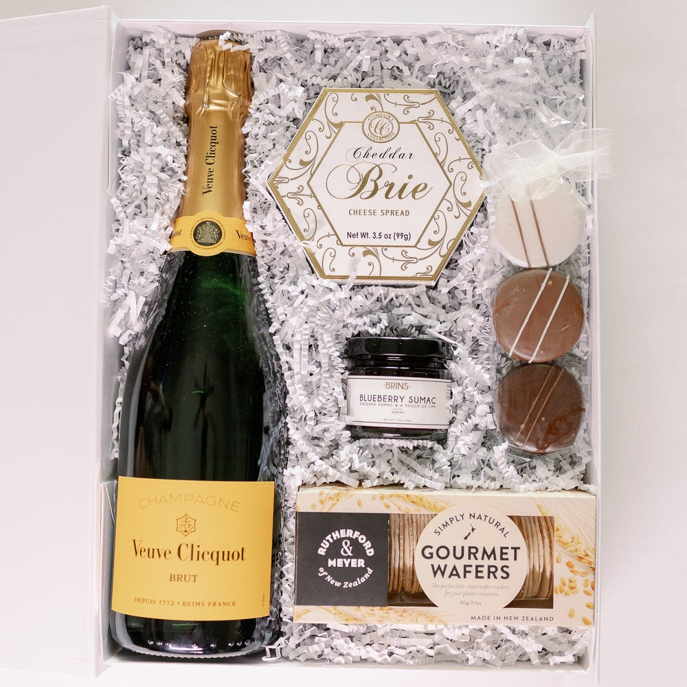 Photograph of a luxury gourmet gift box featuring a bottle of Veuve Clicquot Brut Champagne with its iconic yellow label, a gold-patterned hexagonal box of Cheddar Brie, and Rutherford & Meyer Gourmet Wafers nestled in white shredded paper with three drizzled chocolates and a jar of blueberry sumac spread