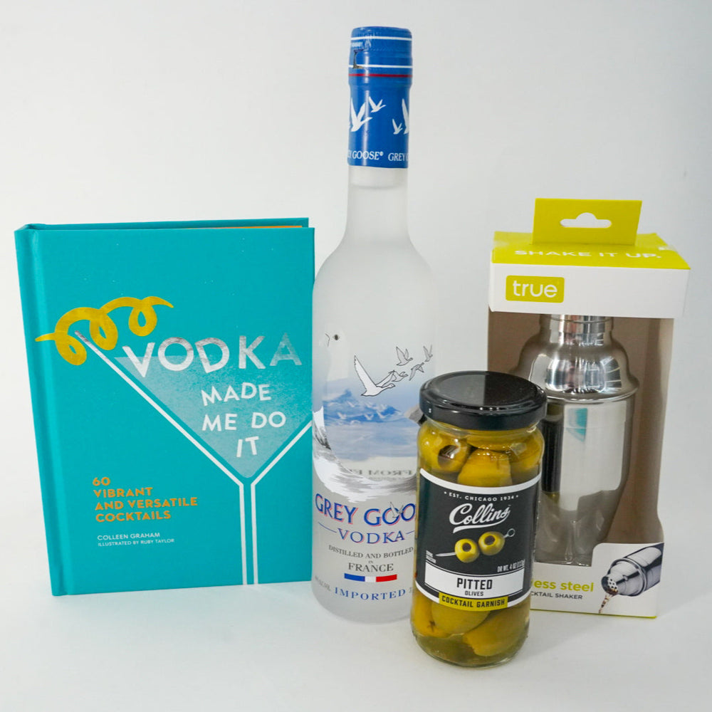 Photograph of a vodka cocktail gift set on a white background featuring the teal book Vodka Made Me Do It by Colleen Graham, a frosted Grey Goose bottle with a blue neck seal, and a stainless steel shaker in a lime yellow box