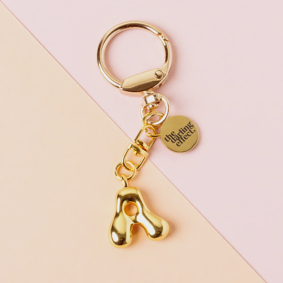 Gold polished metal keychain by The Darling Effect featuring a puffy bubble letter A charm and a signature circular tag on a minimalist diagonal pink and peach background