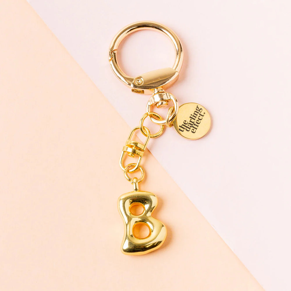 Photograph by the darling effect of a gold bubble initial B keychain featuring a polished 3D letter, a branded circular charm, and a lobster claw clasp on a diagonal two-toned pink background