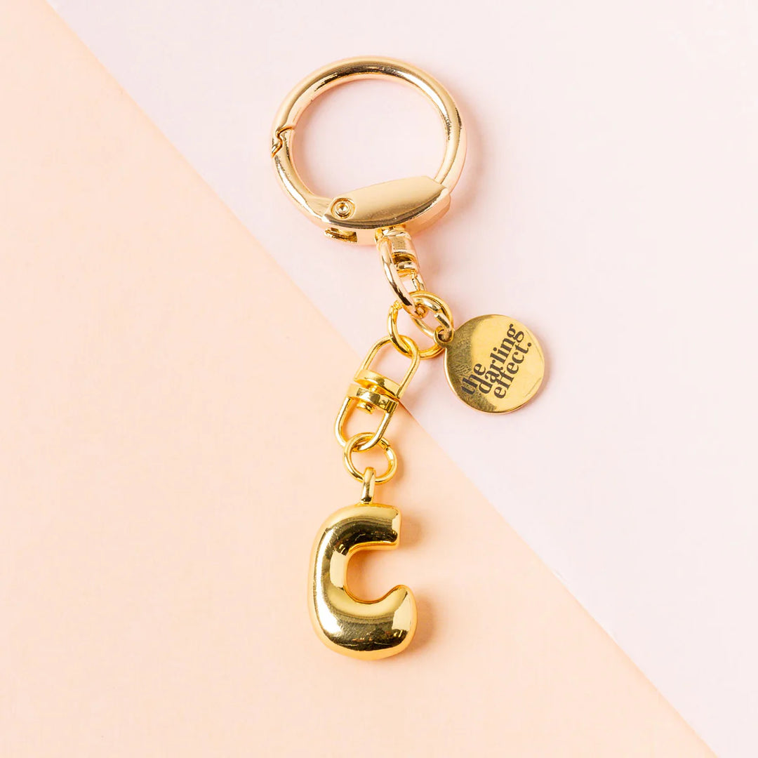 Gold-toned metal keychain by the darling effect featuring a polished 3D bubble initial C, an engraved round logo disc, and a lobster clasp against a diagonal pink and peach background