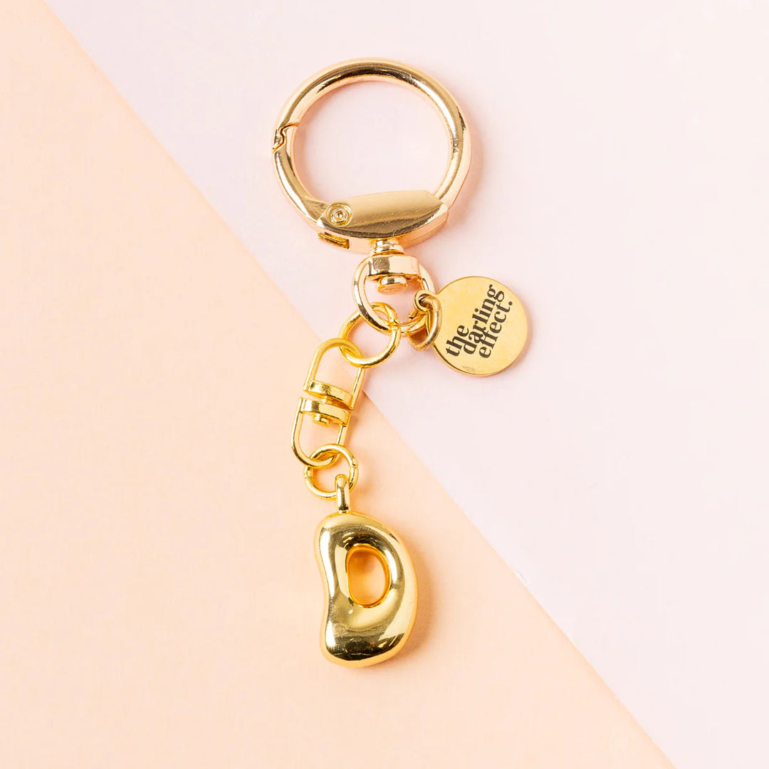 Photograph by the darling effect of a gold-toned metal keychain featuring a chunky puffy letter D charm and a circular logo tag on a diagonal pink and peach background