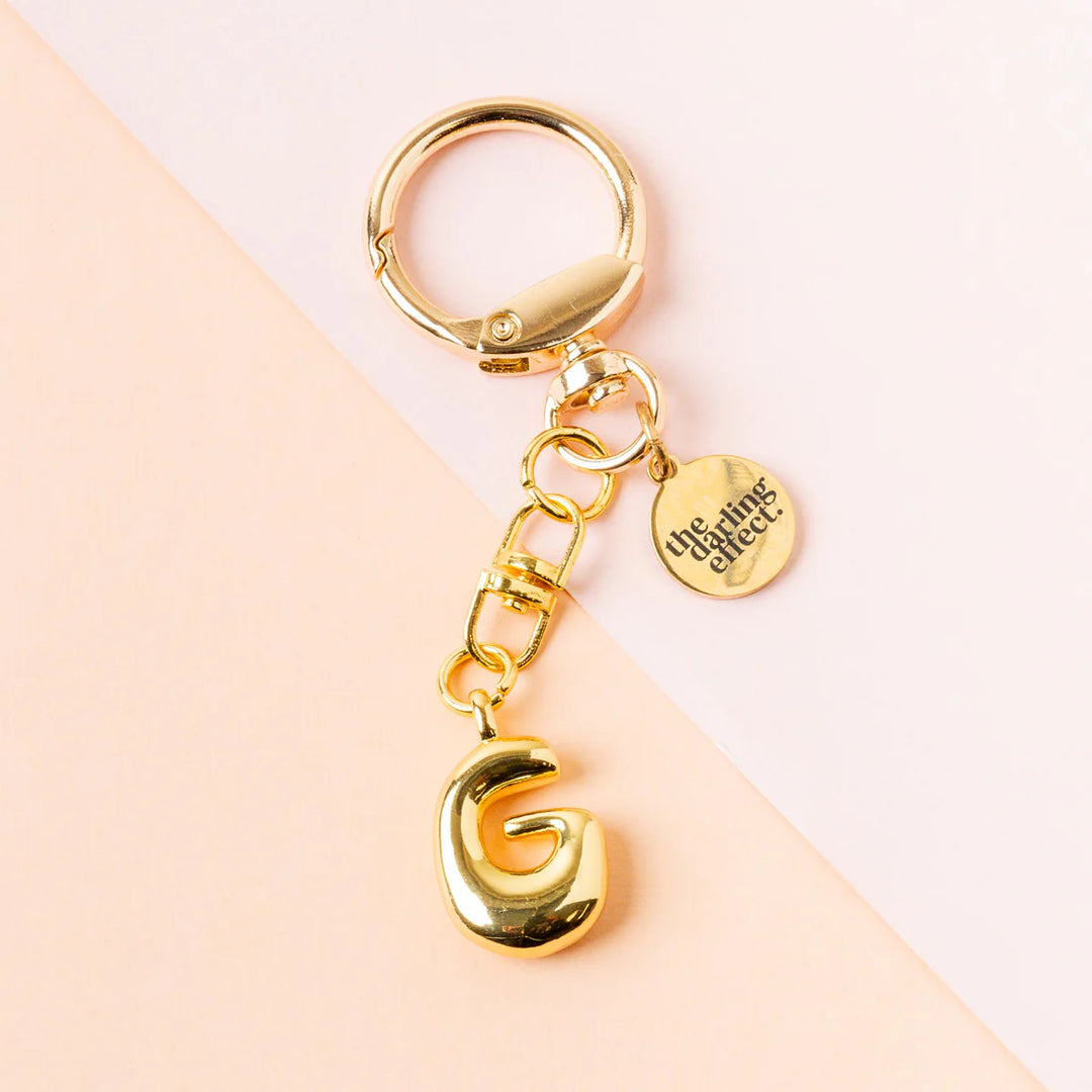 Polished gold metal keychain by The Darling Effect featuring a puffy capital letter G and a circular engraved brand charm set against a diagonal pink and peach background