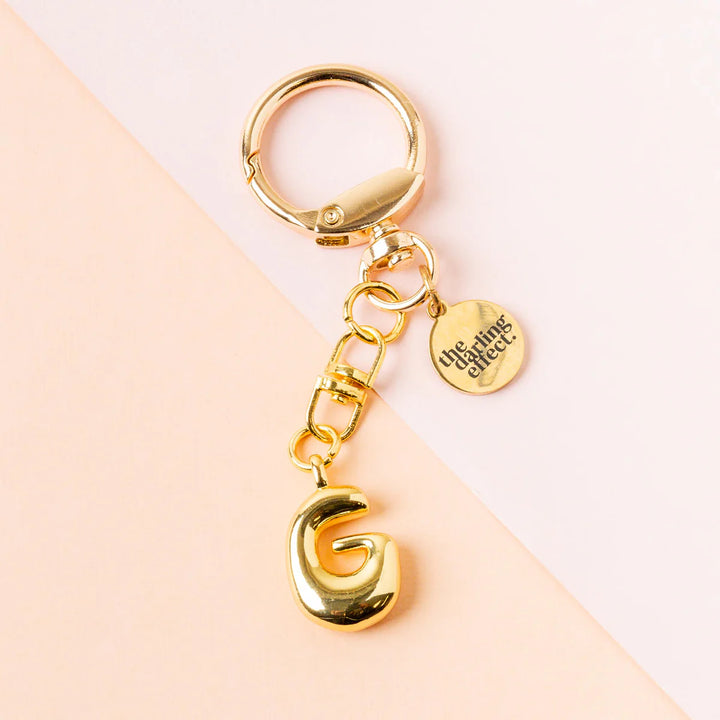 Polished gold metal keychain by The Darling Effect featuring a puffy capital letter G and a circular engraved brand charm set against a diagonal pink and peach background