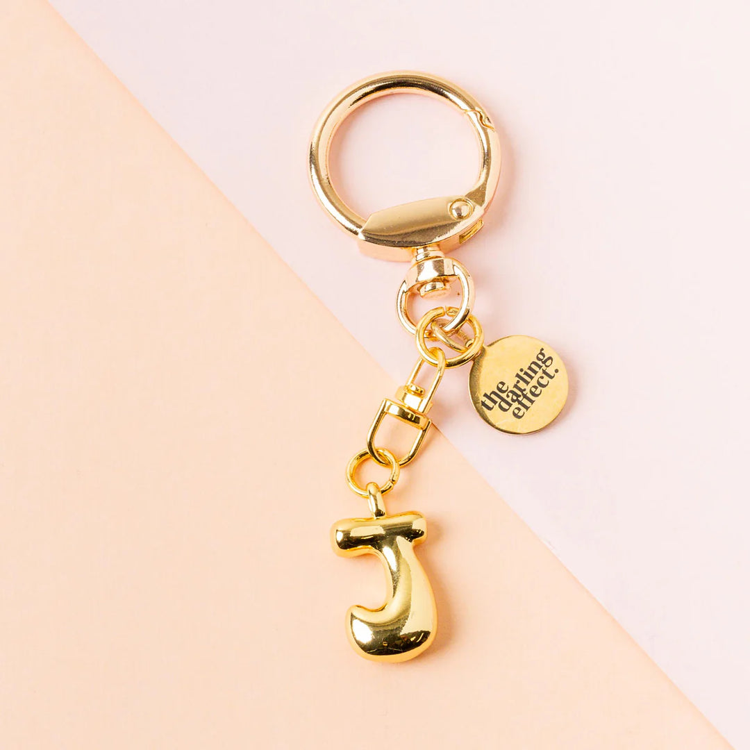 Polished gold metal accessory by The Darling Effect featuring a puffy 3D bubble letter J keychain with an engraved circular brand tag and a large spring-gate clasp on a diagonal peach and pink background