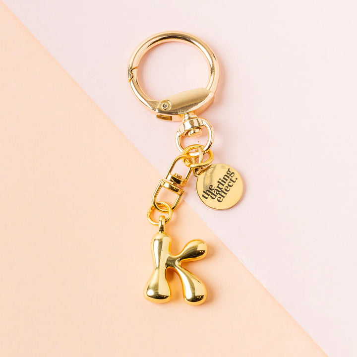Product photograph by The Darling Effect of a gold bubble letter K keychain featuring a high-shine metallic finish and a small engraved logo charm resting on a two-tone pastel pink and peach background