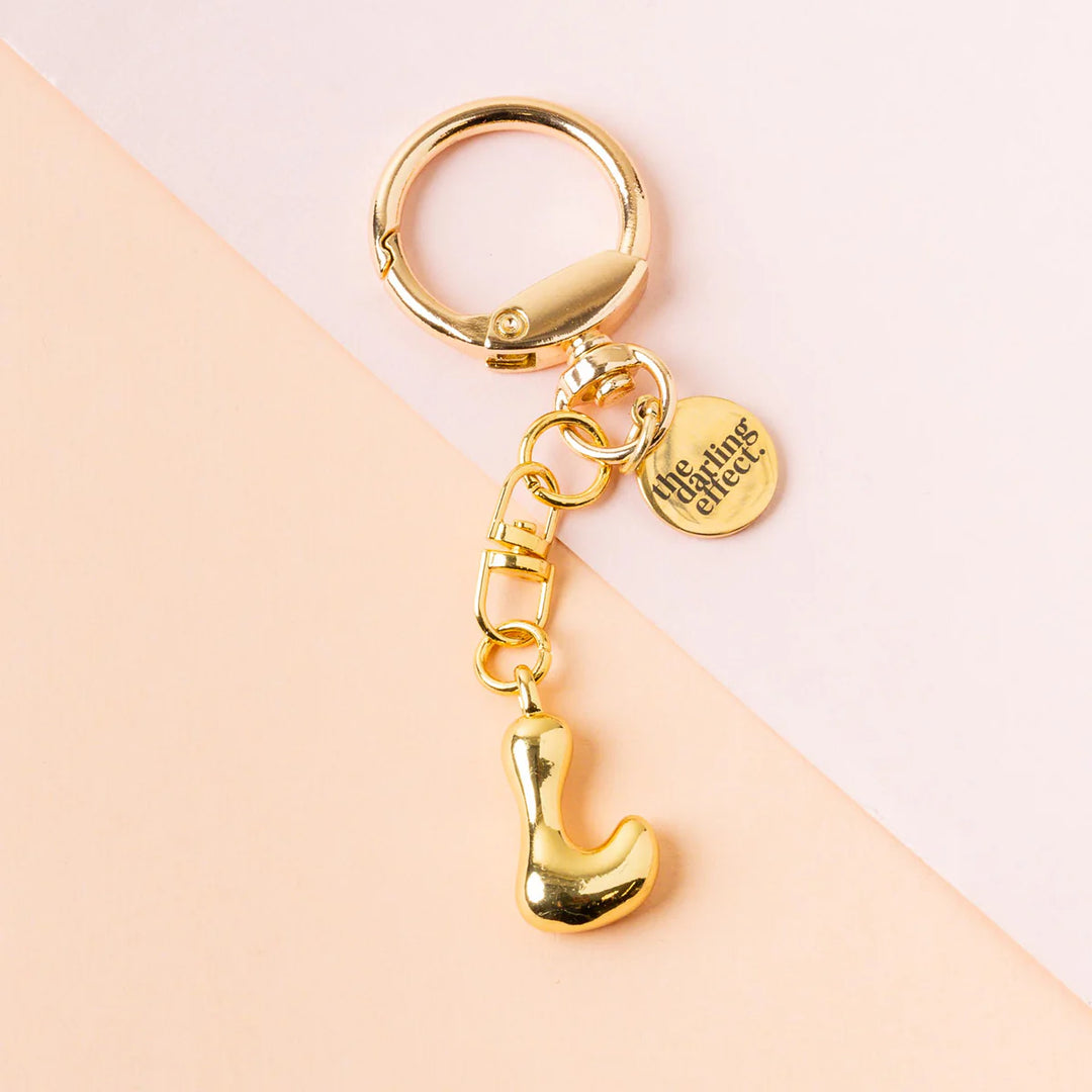 Product photograph by The Darling Effect of a gold initial keychain on a pastel background featuring a polished circular clasp, an engraved logo tag, and a puffy metallic letter L pendant with a high-shine finish