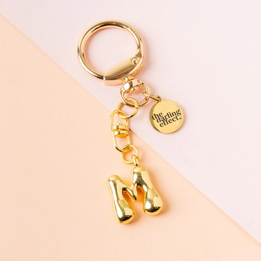 Gold metal keychain by the darling effect featuring a high-shine 3D bubble-letter M and a circular brand charm on a diagonal pastel pink and peach background