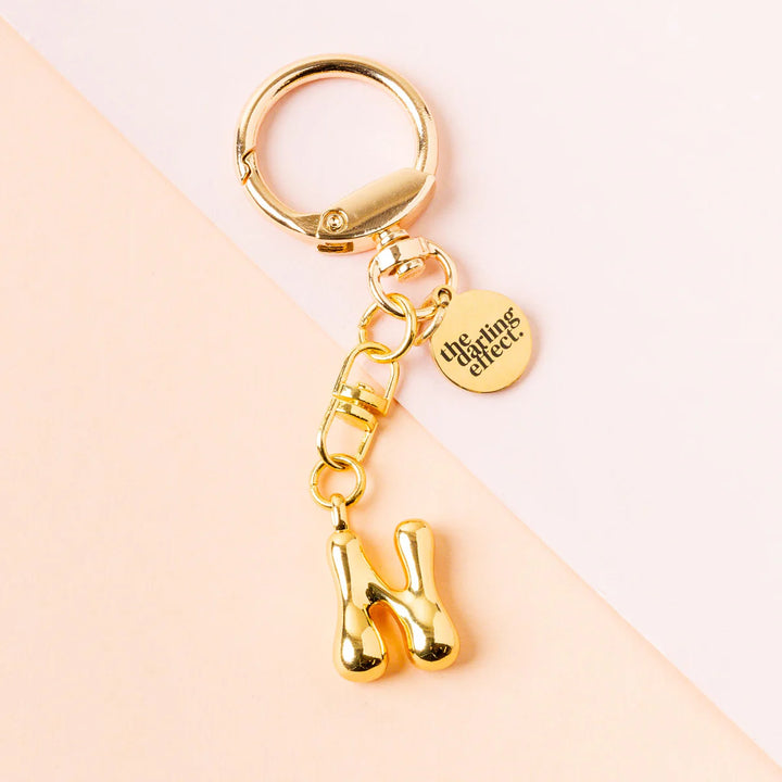 Product photograph by The Darling Effect of a metallic gold initial N keychain featuring a puffy balloon-style letter charm and a circular logo tag set against a diagonal two-tone pink and peach background