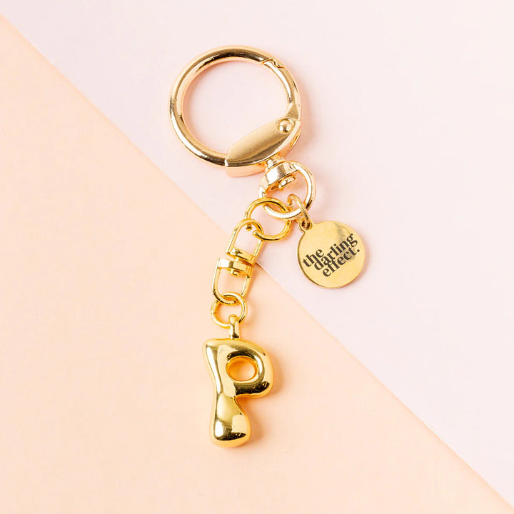 Product photograph by The Darling Effect of a polished gold keychain on a diagonal pink background showcasing a puffy bubble-style letter P charm and a round brand-engraved disk
