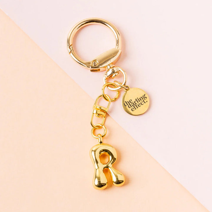 Gold-plated metal keychain by the darling effect featuring a puffy bubble-style letter R charm and an engraved circular logo tag on a split pastel pink and peach background