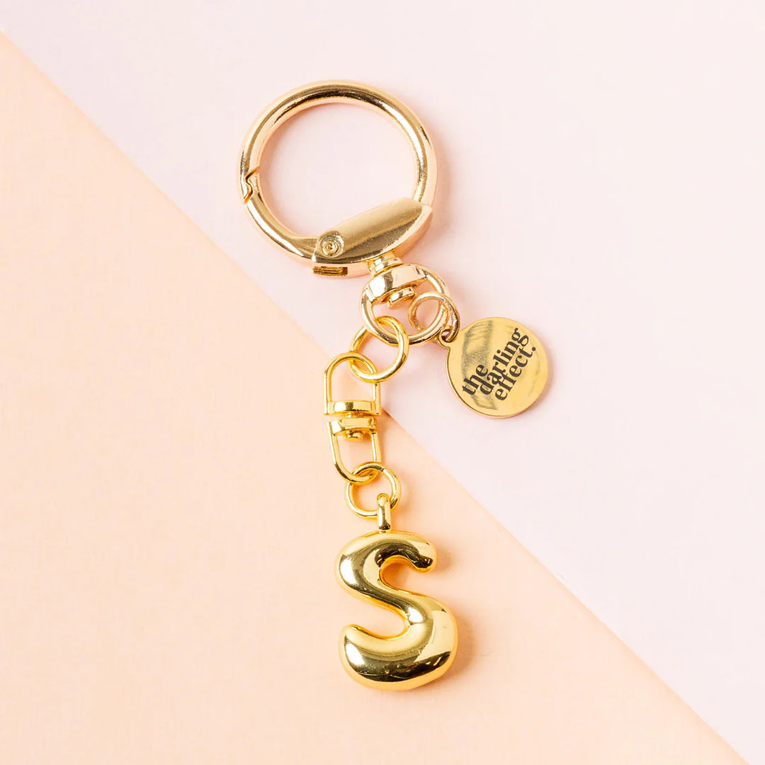 Gold metal keychain by The Darling Effect featuring a polished puffy letter S charm on a diagonal two-tone peach and pink background with a small circular logo tag and a shiny metallic finish