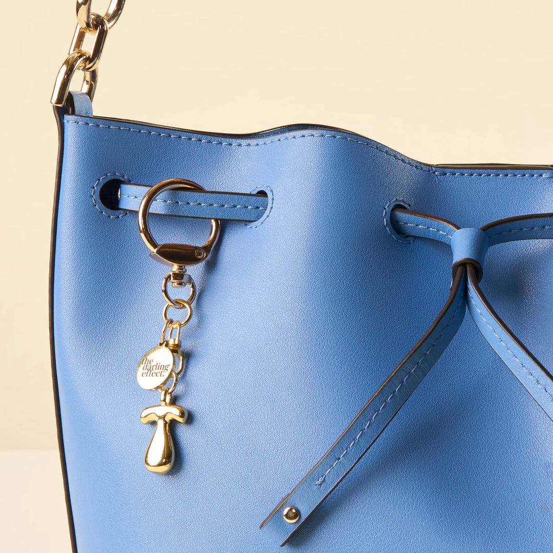 Photograph by The Darling Effect of a cornflower blue pebbled leather handbag featuring a gold chain strap, a polished gold letter T charm, and a circular branded pendant against a light cream background