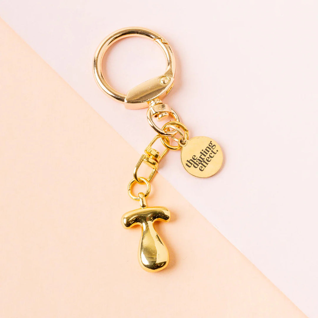 Gold-toned metal keychain by The Darling Effect featuring a polished bubble letter T charm and an engraved circular logo tag resting on a diagonal peach and pink pastel background