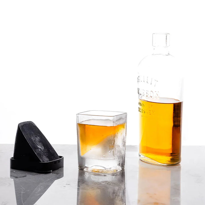 Studio photograph of a Bulleit bourbon bottle and whiskey wedge glass on a reflective surface featuring a large slanted ice block, a square rocks glass filled with golden liquid, and a black silicone mold