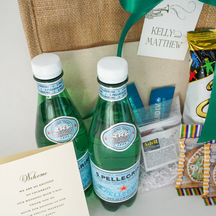 Digital photograph of a wedding welcome gift kit in a burlap bag featuring two green glass San Pellegrino bottles with the iconic Italian spa architecture on the labels alongside a personalized Kelly and Matthew tag tied with a vibrant emerald green ribbon