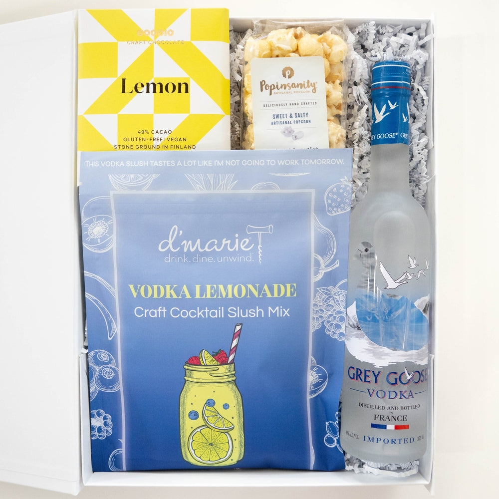 Photograph of a curated cocktail gift box featuring a frosted bottle of Grey Goose Vodka and a d'marie Vodka Lemonade slush mix pouch alongside a bright yellow geometric lemon chocolate bar and artisanal popcorn arranged inside a white box with shredded paper