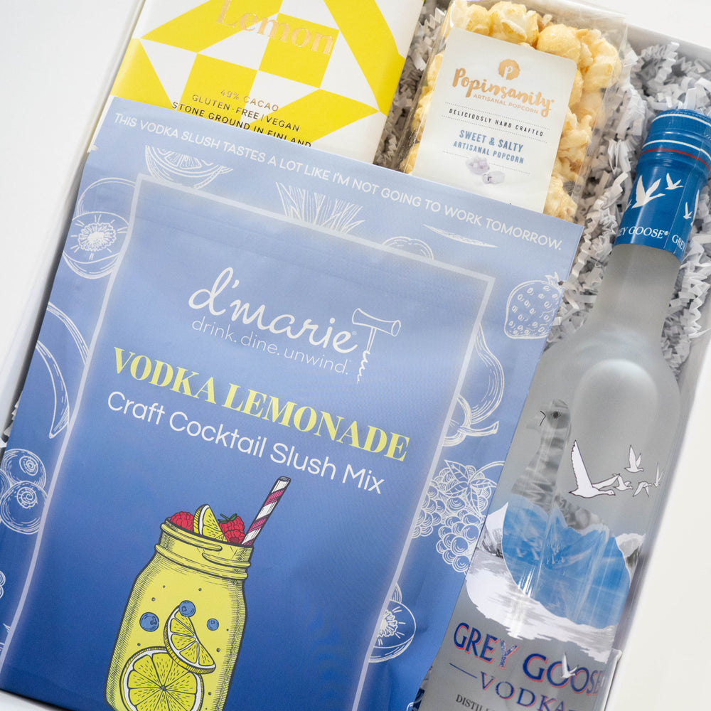 Photograph of a luxury cocktail gift box containing a frosted bottle of Grey Goose Vodka, a d'marie slush mix pouch, and a vibrant yellow geometric lemon chocolate bar resting on white crinkle paper