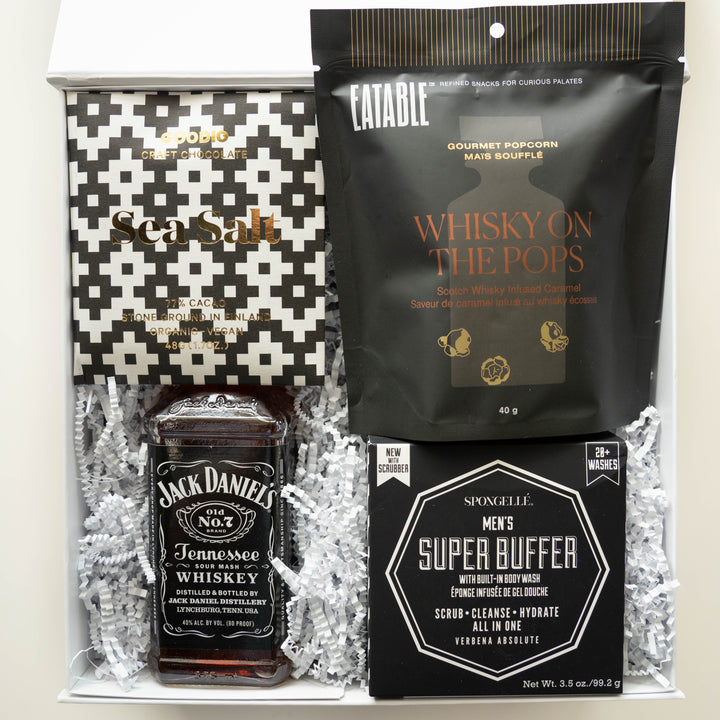 Studio photograph of a luxury gift box featuring a Jack Daniel’s Tennessee Whiskey bottle, a Goodio craft chocolate bar in a black and white geometric wrapper, and a Spongellé Men’s Super Buffer nestled in white crinkle-cut paper