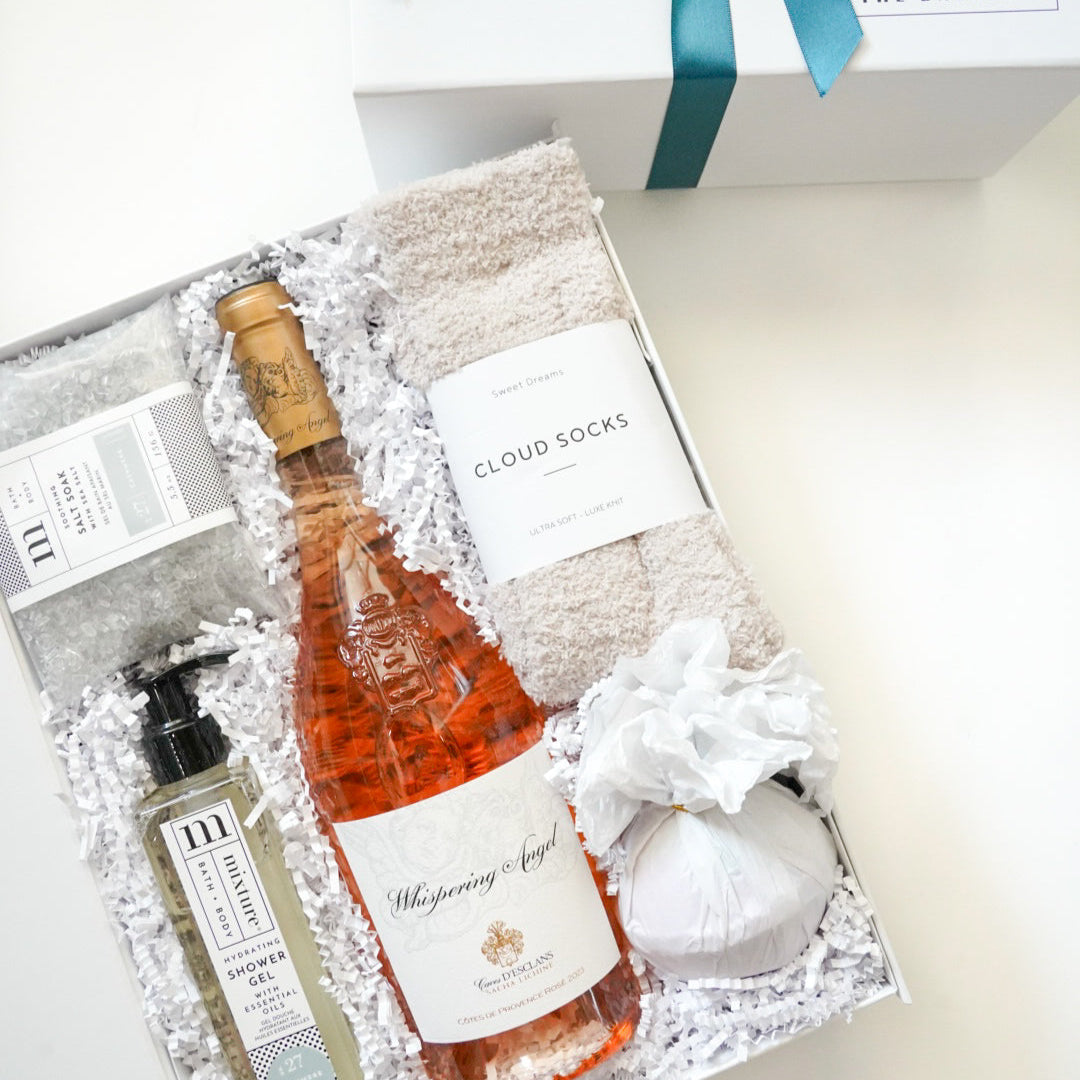 Digital photograph of a luxury self-care gift box featuring a bottle of Whispering Angel Rosé wine, plush cream-colored cloud socks, and a white tissue-wrapped bath bomb nestled in white crinkle paper