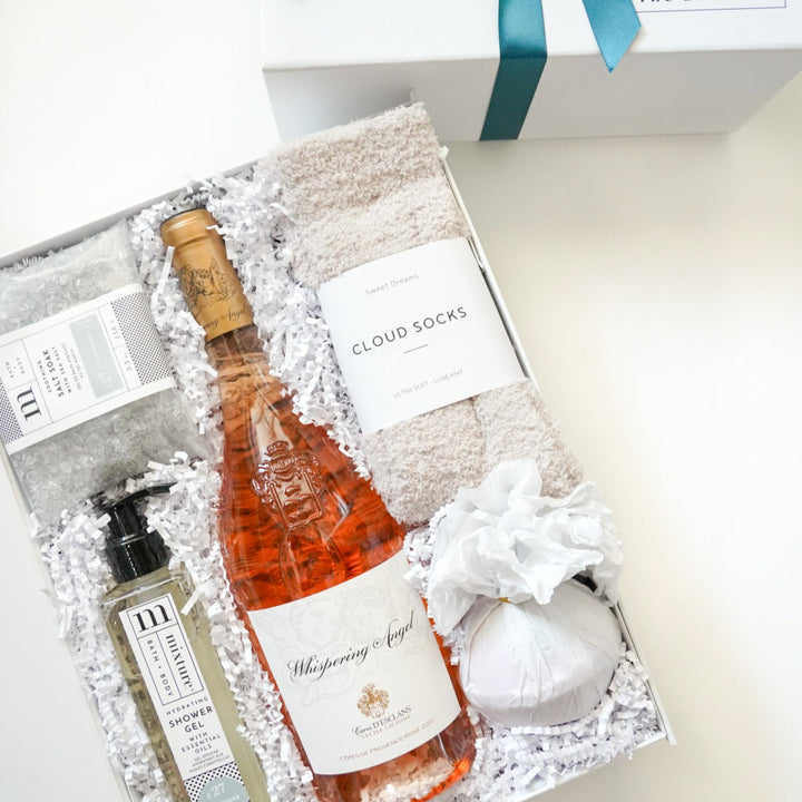 Digital photograph of a luxury self-care gift box featuring a bottle of Whispering Angel Rosé wine, plush cream-colored cloud socks, and a white tissue-wrapped bath bomb nestled in white crinkle paper