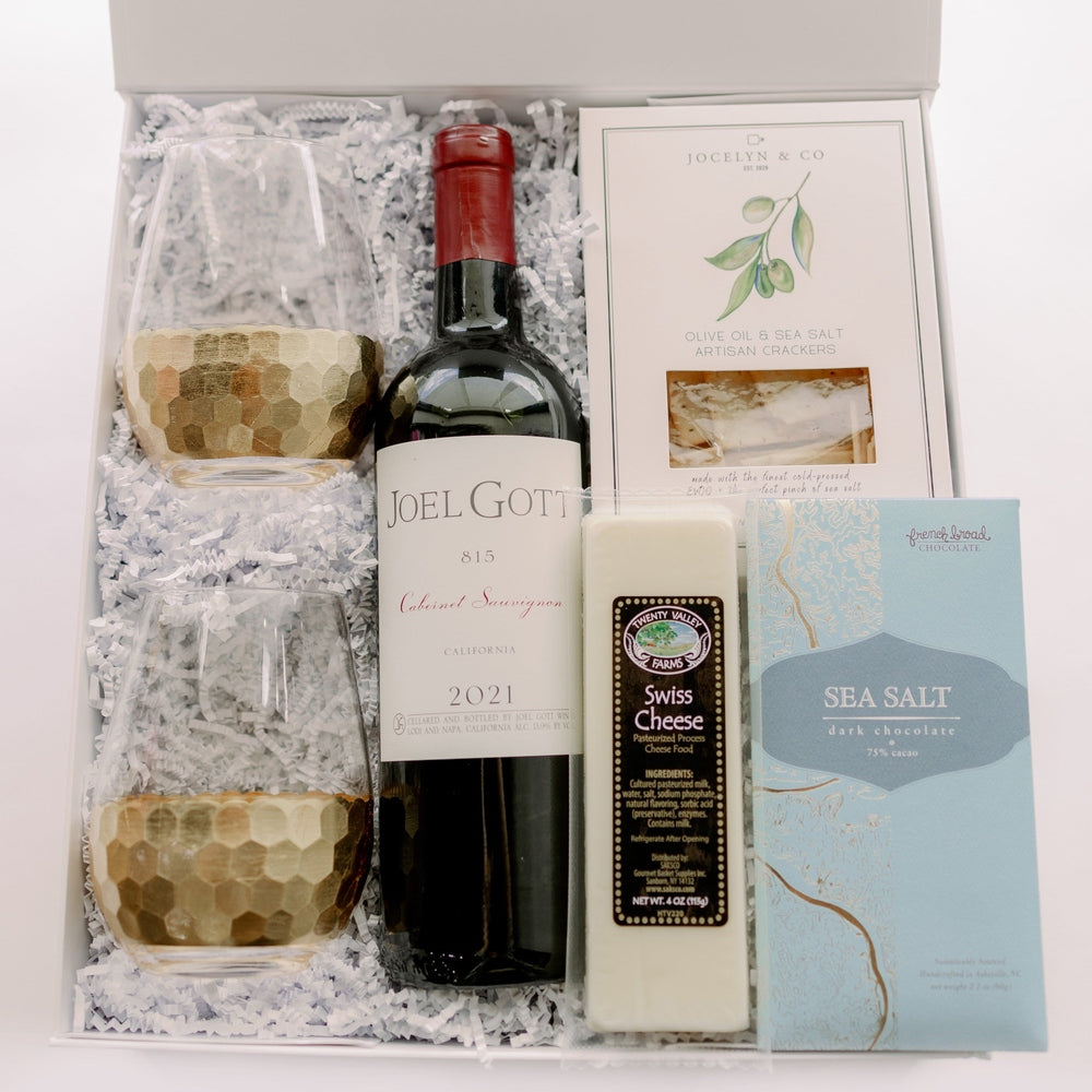 Photograph of a luxury wine gift box featuring a bottle of Joel Gott 815 Cabernet Sauvignon, two stemless glasses with hammered gold honeycomb bases, and a light blue French Broad Sea Salt dark chocolate bar nestled in white crinkle paper