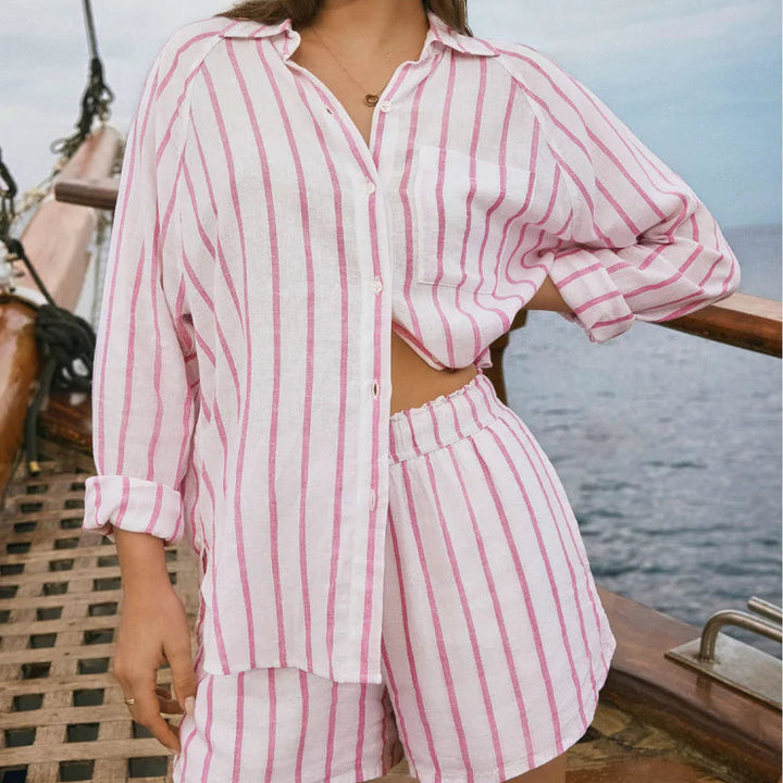 Photograph of a woman wearing a pink and white vertically striped linen shirt and shorts set on a wooden sailboat featuring a front chest pocket and elastic waistband overlooking the open ocean