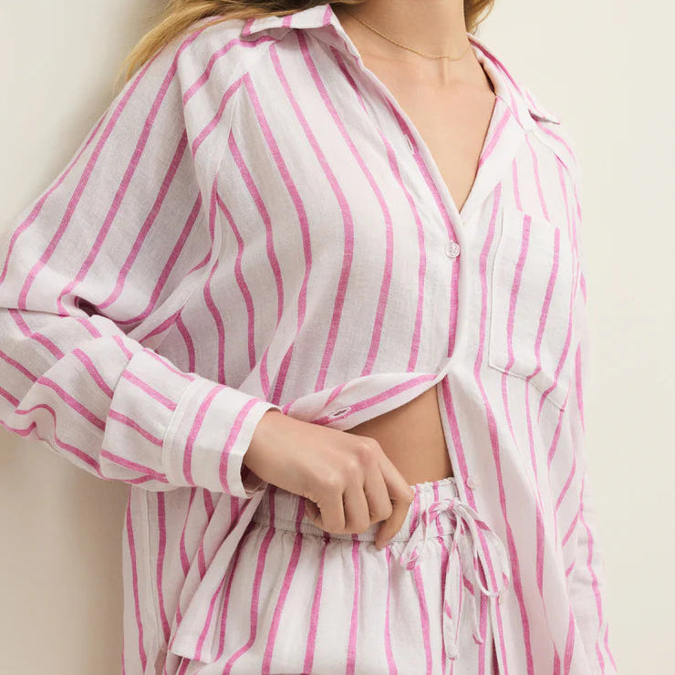 Digital photograph of a woman wearing a pink and white striped linen lounge set in an indoor setting, featuring a button-down shirt with a chest pocket and matching drawstring shorts accented by a delicate gold necklace