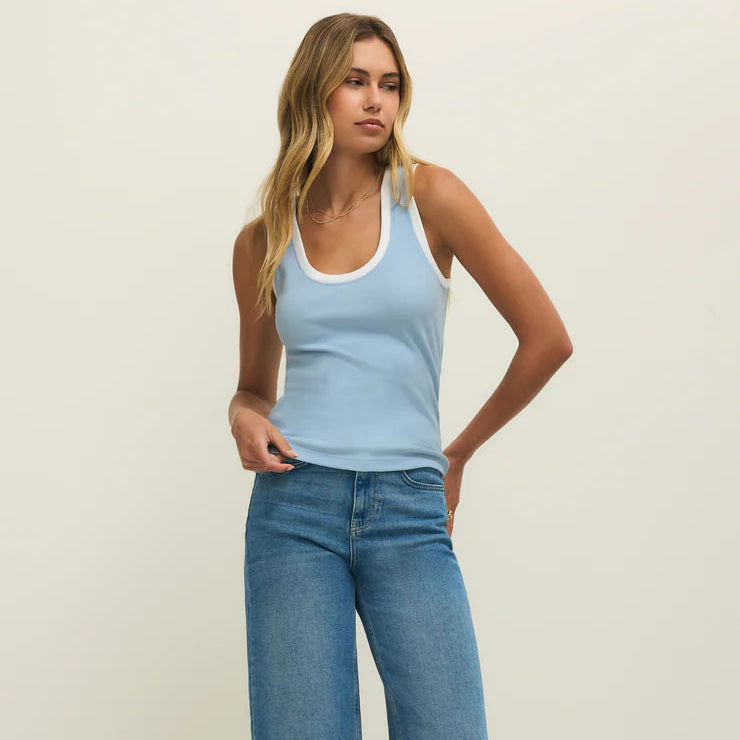 Digital photograph of a female model posing in a minimalist studio wearing a light blue ringer tank top with white trim, high-waisted wide-leg blue jeans, and delicate gold necklaces