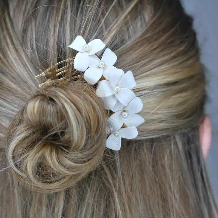 Photograph of a bridal hair accessory featuring five white clay flowers with petite pearl centers pinned into a textured blonde bun showing golden metal prongs