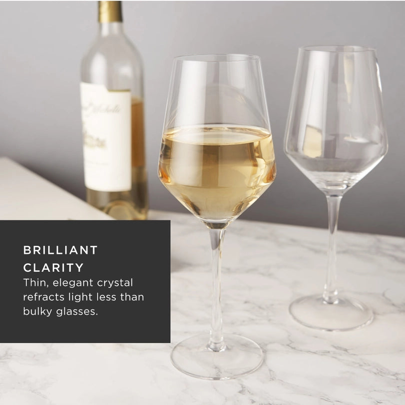 Professional product photograph of two crystal wine glasses on a white marble tabletop, one filled with golden-hued white wine, featuring long slender stems, an angled bowl design, and a blurred Chateau Ste. Michelle wine bottle in the background