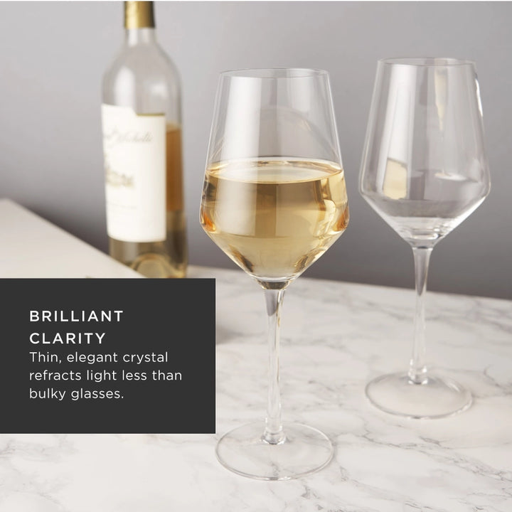 Professional product photograph of two crystal wine glasses on a white marble tabletop, one filled with golden-hued white wine, featuring long slender stems, an angled bowl design, and a blurred Chateau Ste. Michelle wine bottle in the background