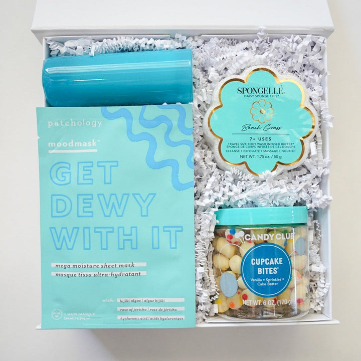 Studio photograph of a curated self-care gift box featuring a teal Patchology sheet mask, a flower-shaped Spongellé body buffer, and a jar of Candy Club cupcake bites on white shredded paper
