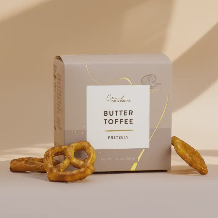 Studio photograph of Grand Provisions Butter Toffee Pretzels featuring a minimalist taupe box with gold foil script and glossy golden-brown glazed pretzels on a neutral beige background with soft shadows