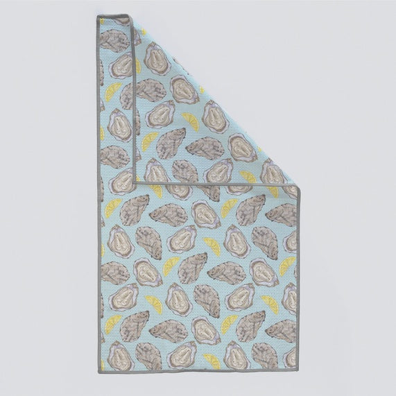 Product photograph of a microfiber kitchen towel featuring a repeating illustrative pattern of textured grey oyster shells and vibrant yellow lemon wedges on a light blue background with a grey stitched border