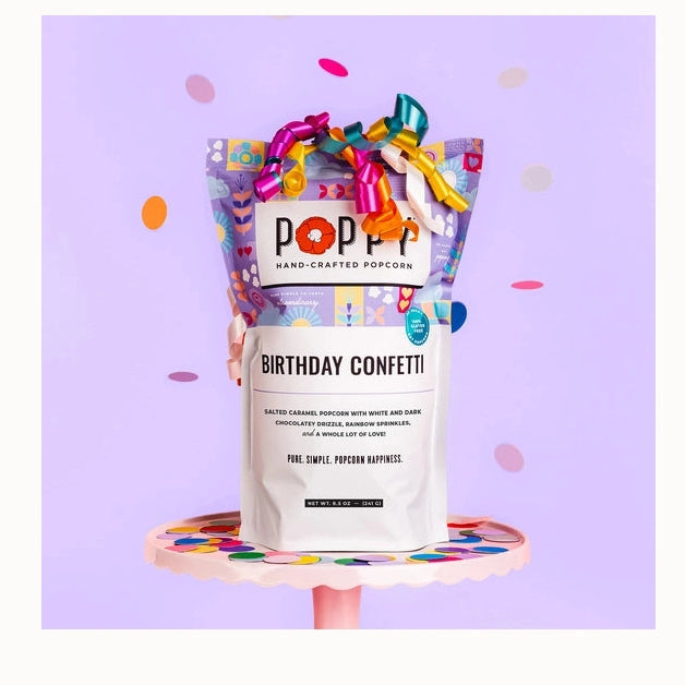 Studio photograph by Poppy Hand-Crafted Popcorn of a Birthday Confetti bag on a pink scalloped cake stand against a lavender background with vibrant curling ribbons and falling multi-colored confetti
