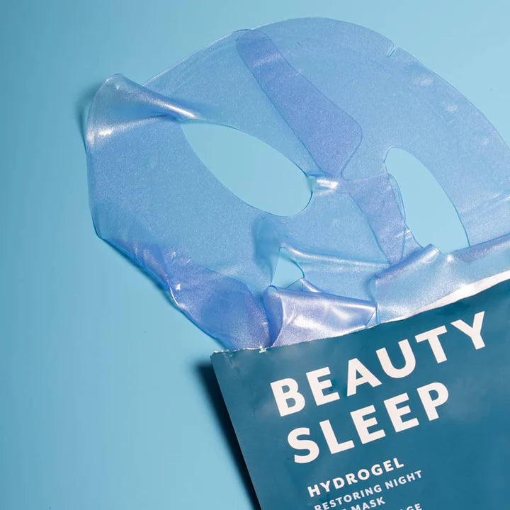 Product photograph of a Beauty Sleep hydrogel face mask on a light blue studio background, highlighting the shimmering translucent blue texture, precision eye cut-outs, and teal packaging with bold white text