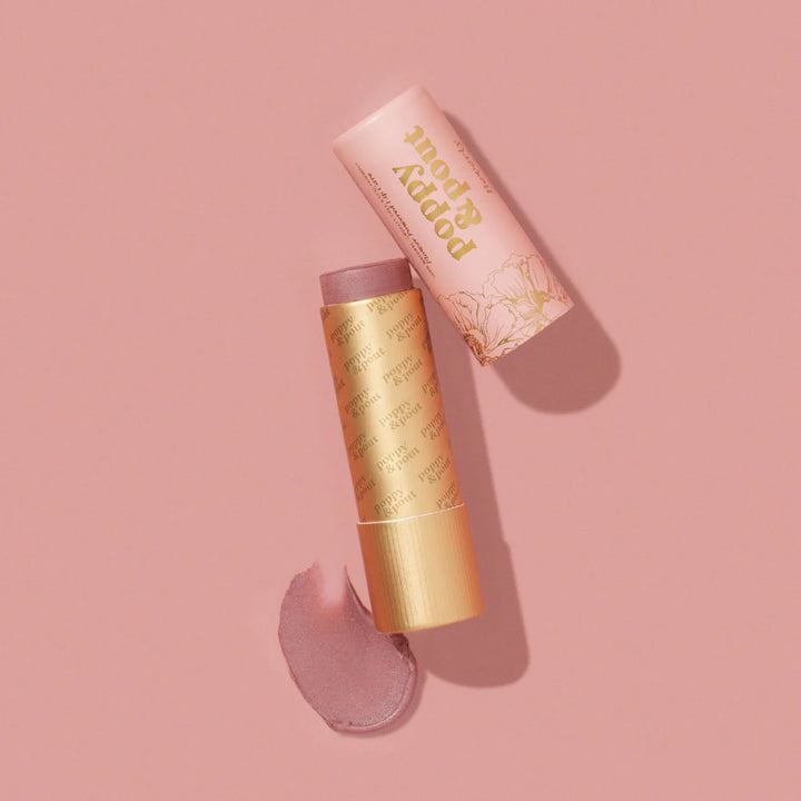 Product photography of Poppy &amp; Pout tinted lip balm on a monochromatic pink surface featuring an open cardboard tube with gold floral illustrations, a shimmery mauve balm stick, and a textured swatch of the product
