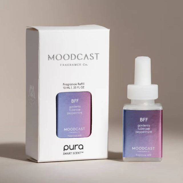 Commercial product photography of Moodcast Fragrance Co. BFF fragrance refill for Pura featuring a white box and glass bottle with a vibrant purple and blue gradient label and scent notes of gardenia, tuberose, and peppermint on a neutral beige surface