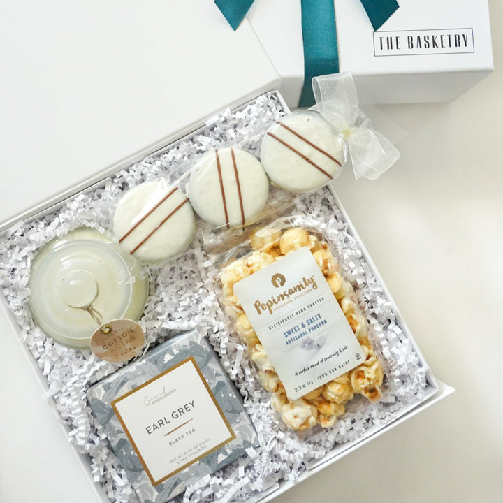 Photograph by The Basketry of a curated gourmet gift box featuring Popinsanity artisanal popcorn, Grand Provisions Earl Grey tea, and three white chocolate-dipped cookies with chocolate stripes nestled in white crinkle paper with a dark teal ribbon