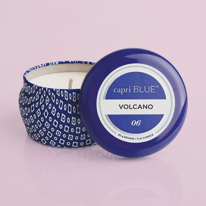 Commercial product photograph by Capri Blue of a Volcano scented travel tin candle featuring a vibrant cobalt blue lid, an intricate blue and white geometric diamond pattern, and white wax on a soft pink background